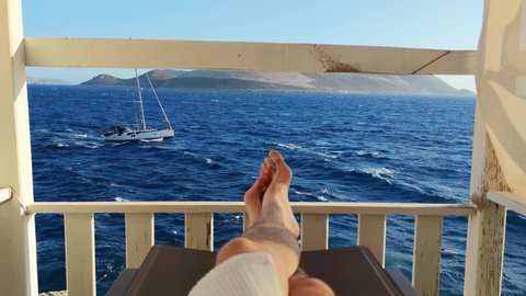Personal perspective of male tourist lying on sunbed in bathrobe and enjoying the seascape and moving ship in slow motion. Low section of a man resting on the pier in front of the ocean and mountains.