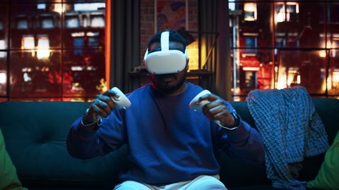 Conceptual Zoom In Shot of a Young Handsome Black Man Putting On VR Headset and Getting Immersed in Virtual Reality. Creative Gamer Sitting on a Sofa at Home, Playing VR Video Games.