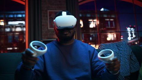 Cinematic Portrait of a Young Stylish Black Man Using Virtual Reality Headset with Joysticks. African American Man Sitting on a Sofa at Home in the Evening, Playing VR Video Games.