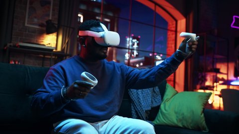 Cinematic Portrait of a Young Stylish Black Man Using Virtual Reality Headset with Controllers. African American Man Sitting on a Sofa at Home in the Evening, Playing VR Video Games.