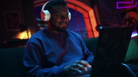 Young Handsome Black Man in Headphones Working from Home on Laptop Computer in Cozy Stylish Loft Apartment. Creative Male Smiling, Checking Social Media, Browsing Internet. Flat with Big Windows.