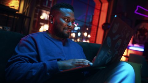 Young Stylish Black Man Working from Home on Laptop Computer with Stickers in Cozy Stylish Loft Apartment. Creative Male Smiling, Checking Social Media, Browsing Internet. Flat with Big Windows.