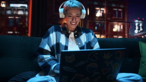 Young Beautiful Woman Wearing Headphones and Working from Home on Laptop Computer in Cozy Stylish Loft Apartment. Creative Female Smiling, Checking Social Media, Browsing Internet.