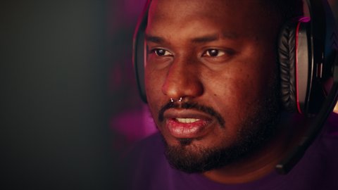 Close Up Portrait of a Black Stylish Young Man with Nose Ring Playing Online Computer Video Game in the Evening at Home. Gamer Discussing Tactics with Teammates while Talking into Headset.