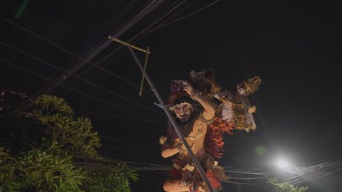  Bali, Indonesia - March, 3, 2022: Unstable live camera view of huge ogo-ogo statue made from paper-mache dragged by crowd of balinese people and hardly fitting in space under electric wires. Night.