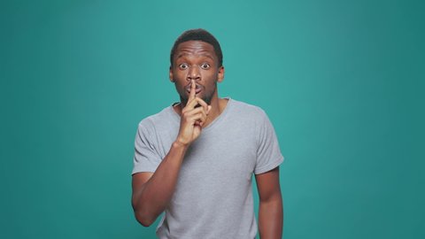 Portrait of secretive man doing hush sign in front of camera, putting index finger over mouth and lips. Male model showing mute and silence gesture, asking for privacy about confidential gossip.