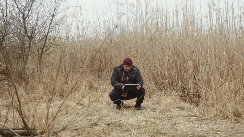 a squatting traveler looks at a tablet with a vintage film camera on his neck against the background of lake reeds.