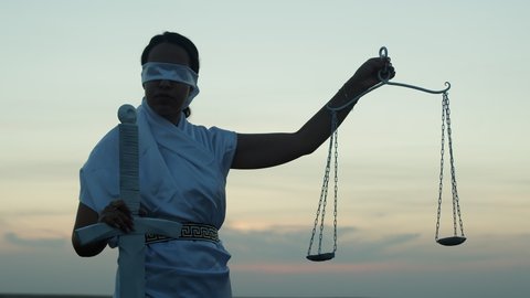 Greek goddess stand against background sunset, hold sword and scales. Woman in white clothes with blindfold turn head to side, lower scales down, outdoor. Concept Greek mythology and legal justice
