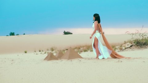Video with noise. young Egyptian woman goddess Cleopatra walks. White antique vintage dress orange cloak gold jewelry. Girl brunette long loose hair sexy back. blue sky sunny sunset ancient pyramids