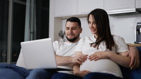 Lovely pregnant woman and man surf the net sitting on the couch with laptop
