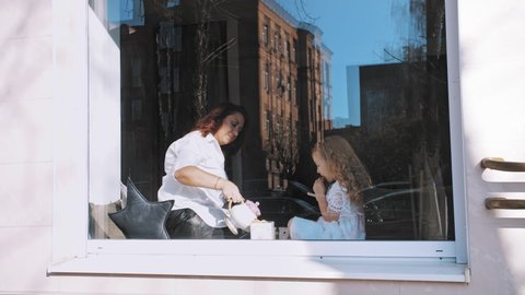 tea party. mother and little daughter are drinking tea with sweets, cookies, sitting by the window, on a large windowsill. view through the window, from outdoors