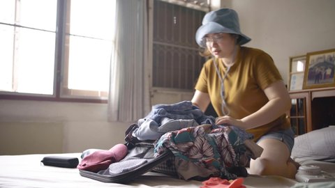 Asian woman try to fit everything in the luggage with struggling, belongings such as sunglasses, polaroid camera, cosmetic bag and small bag. Packing to go on a getaway vacation. Slow-motion video.