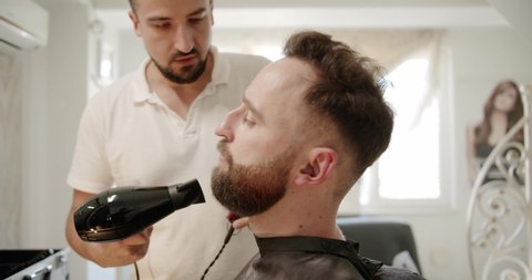 An attractive man with a modern haircut. The hairdresser works with a hair dryer and combs a beard. Hairdresser's. Hairdresser and hairdresser in a retro hair salon. Men's hairstyle.