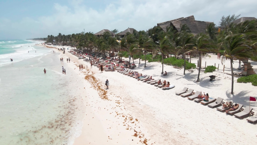 Aerial drone shot over beach in Tulum, Mexico. People chilling, laying around, swimming. Resorts and hotels in the background. Sunny and windy.