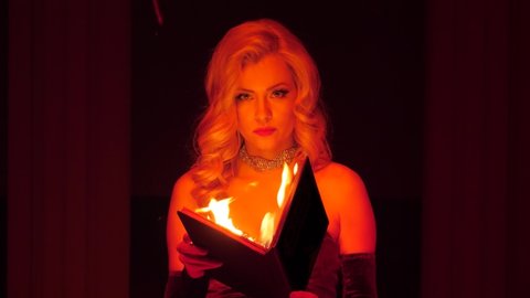 Beautiful woman in dress holds burning old book in her hands on dark background at night. Glamor luxury lady with magic fire book looking at camera. Mysterious ritual concept