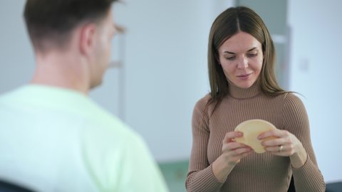 Portrait of young Caucasian beautiful woman choosing implants for breast augmentation in clinic indoors. Positive slim lady talking with expert plastic surgeon approving choice smiling
