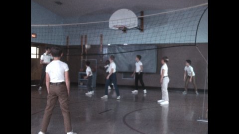 1960s: Young boys play volleyball inside gymnasium. Woman holds band-aid near cut on finger of child.