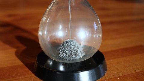 An hourglass made of yellow metal shavings passes through a funnel, symbolizing the concept of time in motion. A clock on a wooden table.