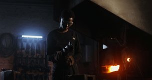 A young blacksmith of African American appearance in goggles and an apron cuts off part of a metal blank using an angle grinder, metal sparks fly in different directions. video in 4k, red komodo