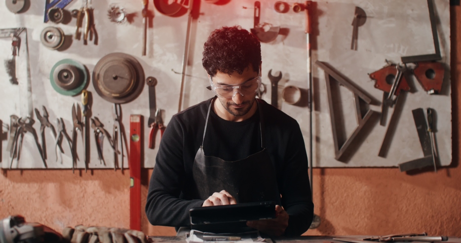 A young blacksmith in working clothes smiling looking at the camera distracted from work at the tablet, sitting at the work table in the forge. video in 4k, red komodo