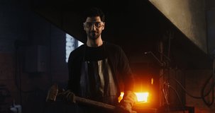 A young bearded man in goggles and a leather apron with a sledgehammer in his hands smiles looking directly at the camera, standing in a modern workshop near a kindled stove. video in 4k, red komodo