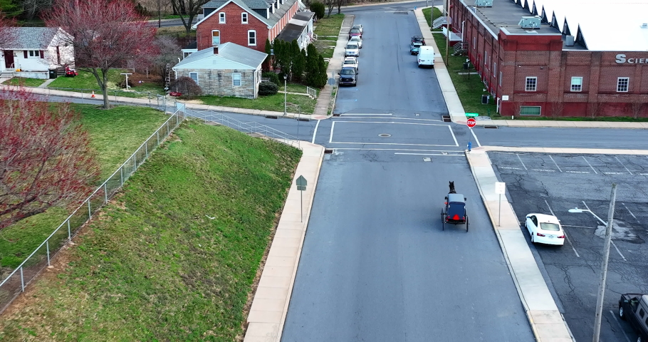 Horse pulls Amish buggy carriage through small town in winter. Homes and businesses line street. Old fashioned antiquated transportation in USA.