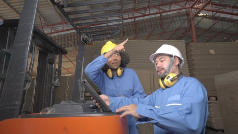 Two warehouse workers, young African female and Asian male, in blue uniforms and helmets with earmuffs hanging on neck, discussing a stocktaking report on a tablet while working on a forklift truck.