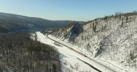 Freight long train carries with cargo carriages in hard to reach wild mountains landscape through a difficult part of Trans Siberian railways. Aerial drone view at winter sunny day.