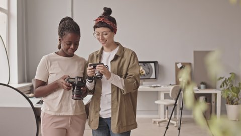 Medium portrait of two young emale photographers with cameras in hands discussing photos on display screen then smiling at camera, standing in bright studio
