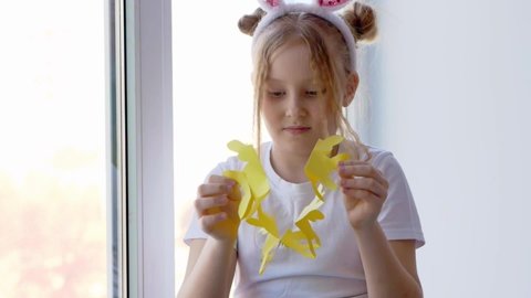 A petite blonde with freckles on her face and bunny ears unrolls and reveals a garland of bunnies cut out of yellow paper. Easter concept for DIY home decor.