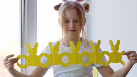 A little blonde girl with freckles on her face and bunny ears smiles and holds and shows a garland of rabbits cut out of yellow paper. Easter concept for DIY home decor.