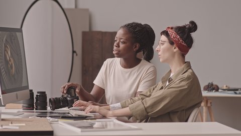 Medium shot of two young multiethnic female photographers having conversation while editing photos on computer sitting at desk in bright photo studio