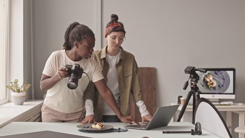 Medium shot of two young multiethnic female photographers working together in studio, taking food pictures on camera and discussing result