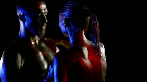 close-up of a couple with golden metallic skin on a black background. they touch each other. the woman is visible from the back. red, blue and white light. the dark key