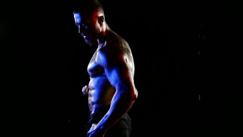 a man with a muscular naked torso and golden-metallic skin poses under water drops on a black background. red, blue and white light. dark key
