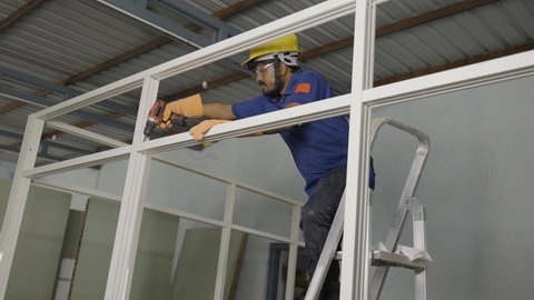 worker fixing screw using drilling machine or gun on aluminium partition frame at construction site - concept of maintenance service, blue collar jobs and safety.