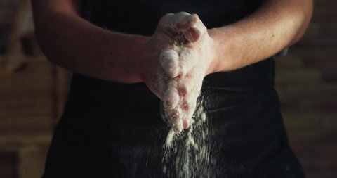 Cinematic macro shot of professional baker chef clapping his hands in dust with flour powder while kneading dough for preparation of pasta, pizza and other pastries in rustic bakery kitchen.