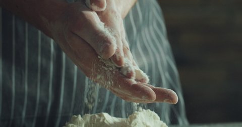 Cinematic macro shot of professional artisan baker chef clapping his hands in dust with flour powder while kneading dough for preparation of pasta, pizza and other pastries in rustic bakery kitchen.