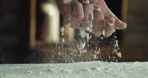 Cinematic macro shot of professional artisan baker chef sprinkles flour dust powder on table while kneading dough for preparation of pasta, pizza and other pastries in rustic bakery kitchen.