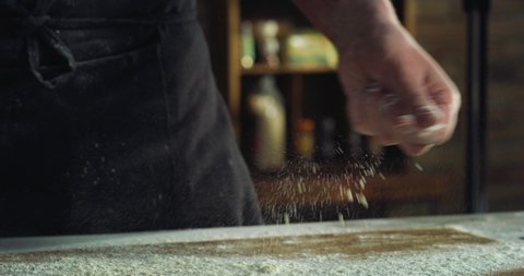 Cinematic macro shot of professional baker chef sprinkles flour dust powder on table while kneading dough for preparation of pasta, pizza and other pastries in rustic bakery kitchen.