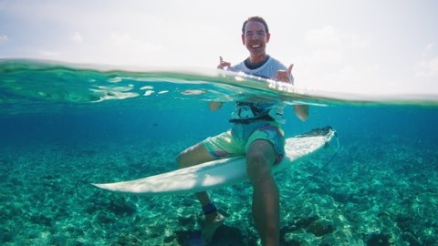 Adult man surfer sits on the surf board in the tropical ocean and shows shaka sign