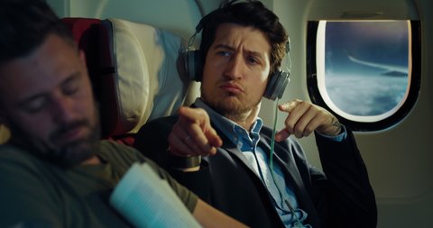 Cinematic shot of young businessman dances crazy while listening music and excuses for disturbing passenger sleeping near in aircraft while traveling with comfort in business class cabin during flight