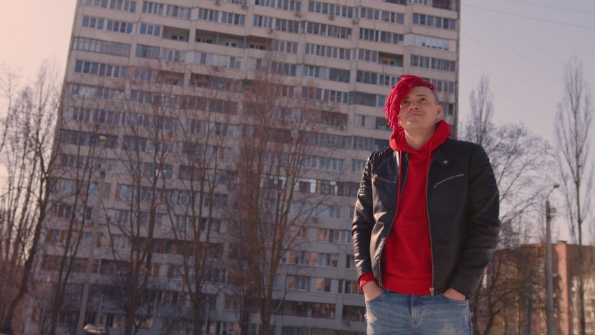 Stylish man in trendy outfit and with red dreadlocks standing on street. Low angle side of young male in a black leather jacket on street and looking away