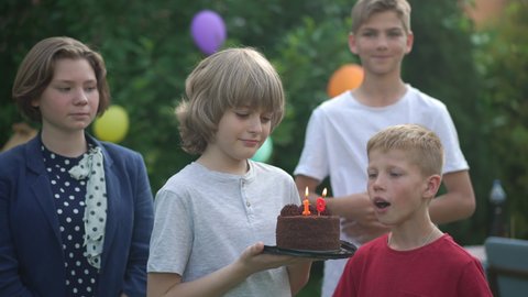 Portrait of handsome Caucasian boy blowing out candle on birthday party with friends congratulating clapping in slow motion. Smiling positive child celebrating outdoors on backyard
