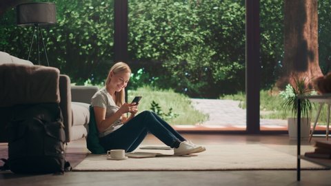 Portrait of Beautiful Young Adult Female with Blond Hair Sitting on a Floor and Using Smartphone in Sunny Living Room. Woman Checking Social Media and Connecting with Friends.