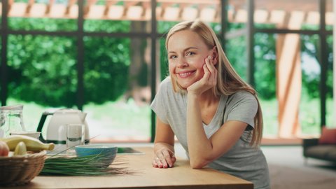 Portrait of Attractive Young Adult Woman with Blond Hair Wearing Gray V-Neck T-Shirt, Leaning on a Table and Smiling Charmingly. Successful Woman Enjoying Leisure Time at Home in Bright Living Room.