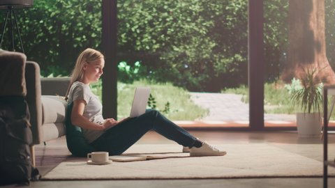 Portrait of Beautiful Young Adult Female with Blond Hair Sitting on a Floor and Using Laptop Computer in Sunny Living Room. Successful Woman Working from Home in Bright Apartment.