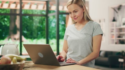 Portrait of Beautiful Young Adult Woman with Blond Hair Wearing Gray V-Neck T-Shirt, Using Laptop Computer while Standing in Living Room. Successful Woman Working from Home in Bright Studio.