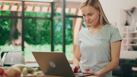 Portrait of Beautiful Young Adult Woman with Blond Hair Wearing Gray V-Neck T-Shirt, Using Laptop Computer while Standing in Living Room. Successful Woman Working from Home in Bright House.