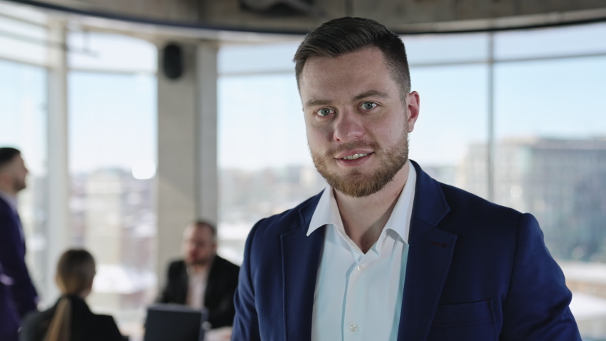 Handsome adult young businessman in dark blue jacket looking straight at camera. Portrait of a modern business owner. Team communicating at backdrop. Royalty-Free Stock Footage #1088578801